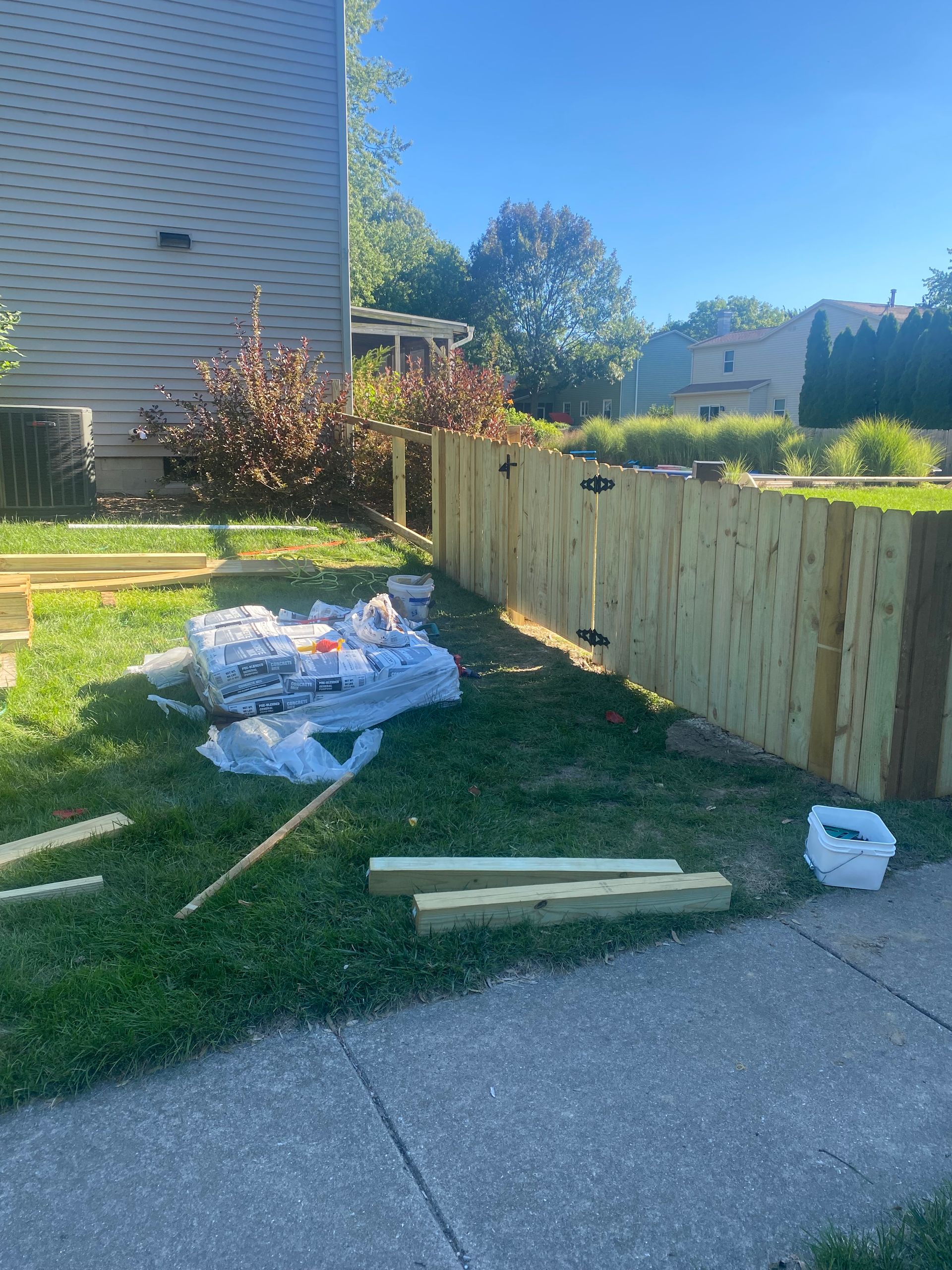 Wooden fence materials and packing debris beside a sidewalk and grassy lawn on a sunny day