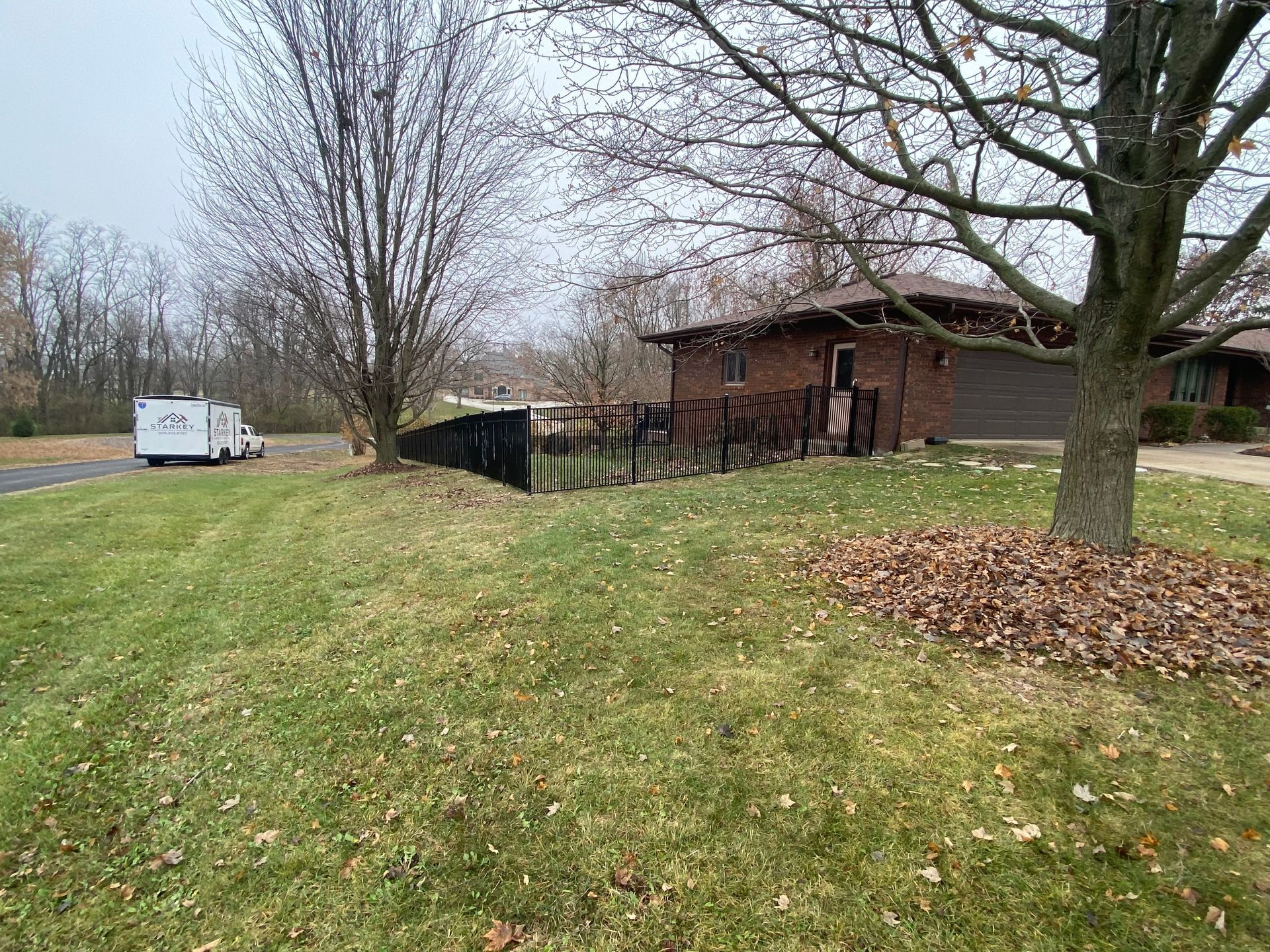 Grassy yard with bare trees, a brown house, and a white trailer on an overcast day