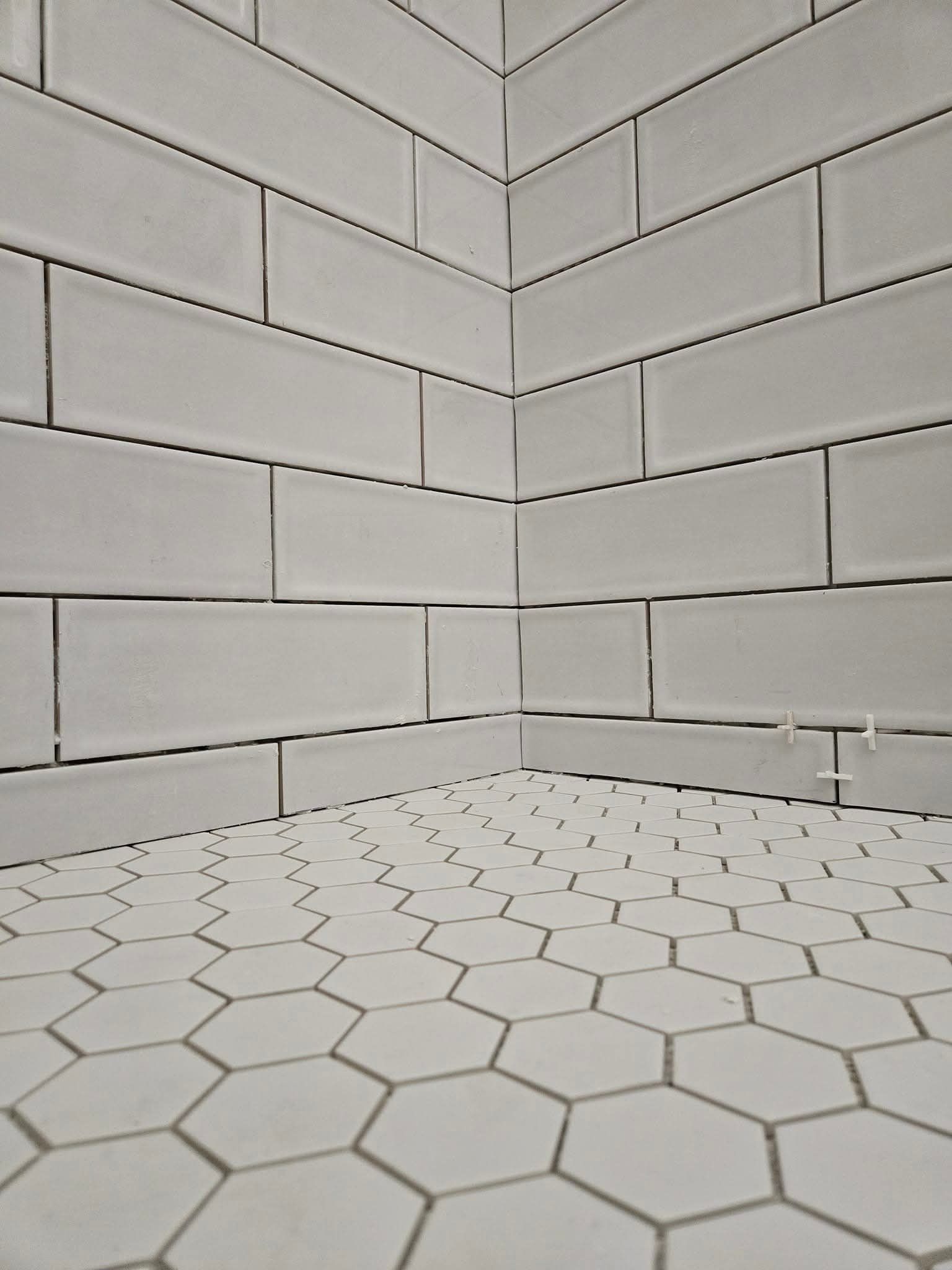 White tiled shower corner, subway tile walls, hexagonal floor tiles, gray grout.