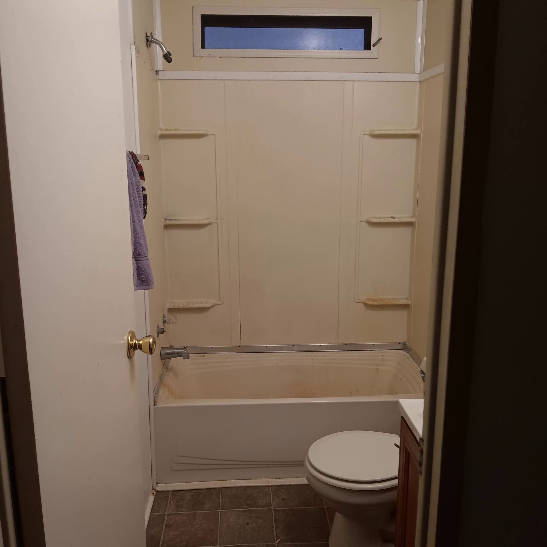 Bathroom interior with a bathtub, toilet, and showerhead. Beige walls, small window, and brown tiled floor.
