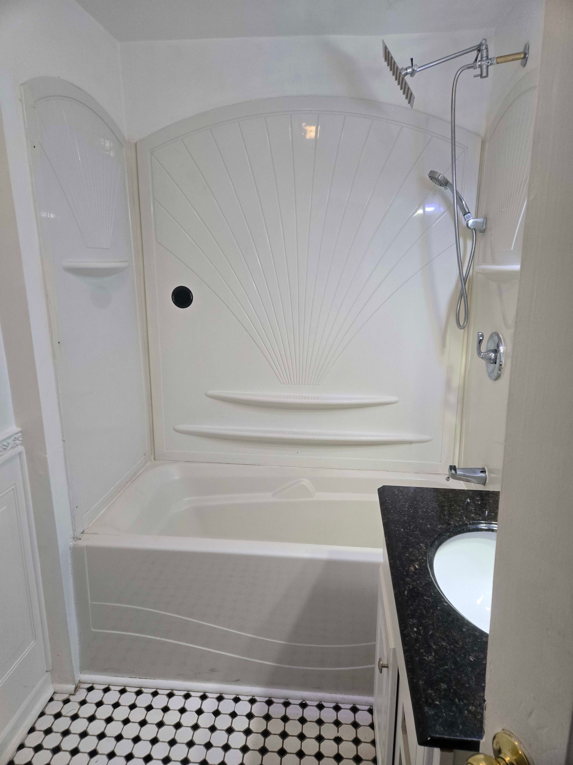 Small bathroom with white tub, shower surround, and black countertop. Black and white tiled floor.