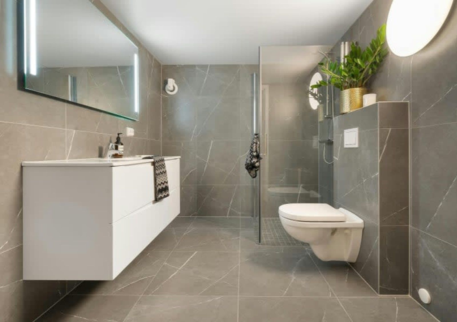 Modern bathroom with gray tile, white vanity, glass shower, and wall-mounted toilet.