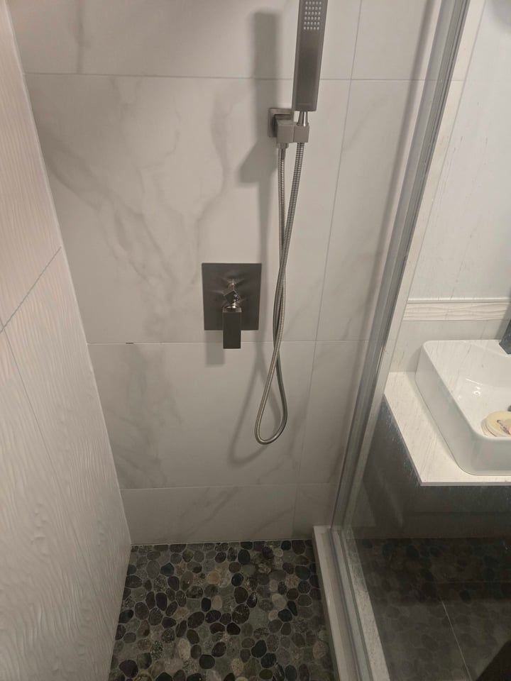 Shower with pebble floor, marble-look tiled walls, and brushed metal shower fixtures. A clear glass shower door is visible.