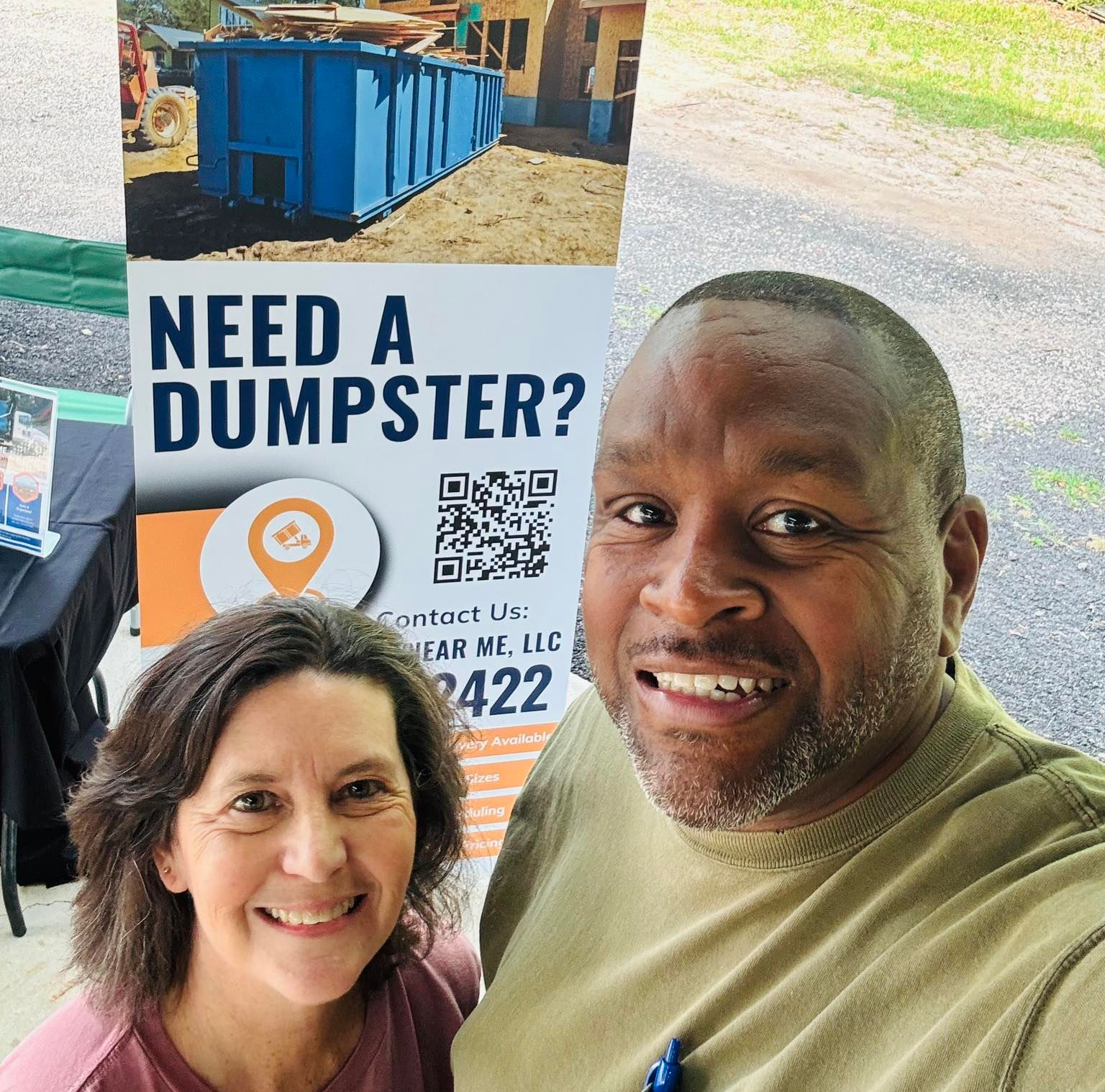 Two people smiling in front of a dumpster rental sign reading “Need a Dumpster?”