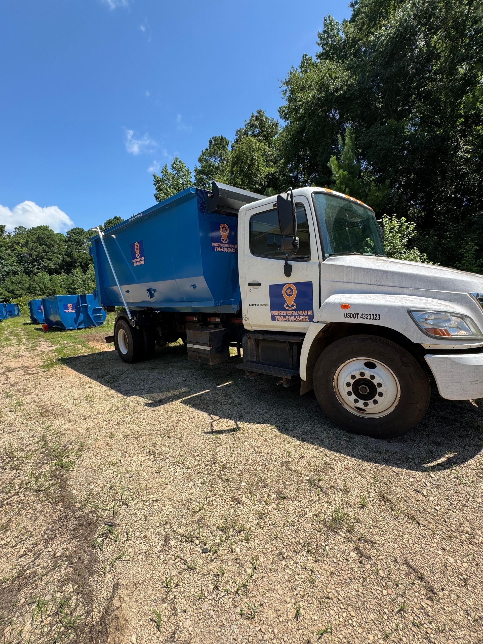 White dump truck with a blue bed driving on a road beside autumn trees