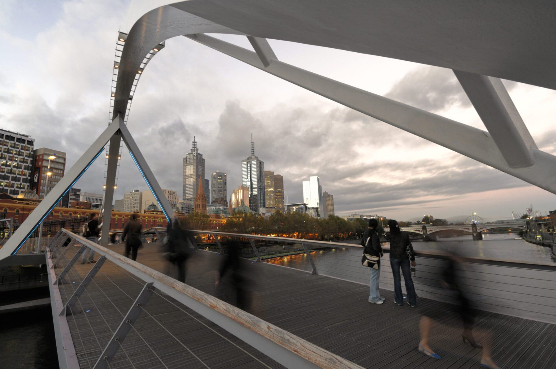 Bridge over water with city skyline in the background, people walking, overcast sky.