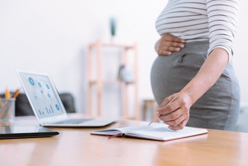Pregnant person at desk, holding a pen over a notebook. Laptop and other office items are also visible.
