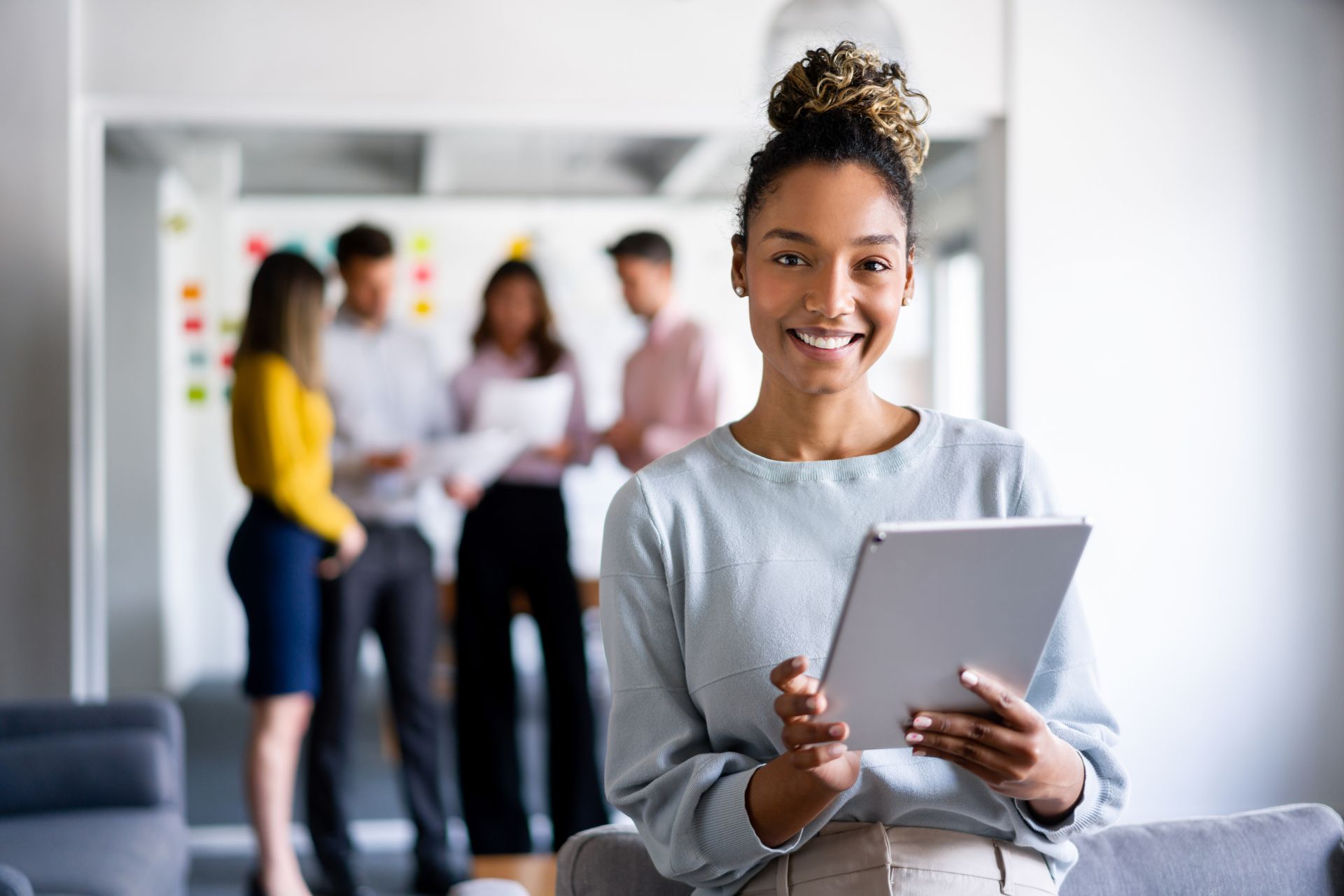 Woman with tablet smiles in office; team in background.