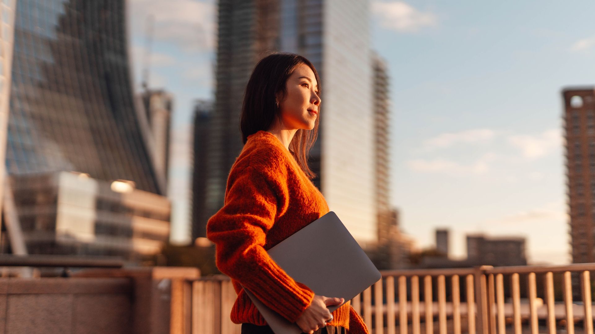 Woman in an orange sweater walking on a city bridge at sunset, holding a gray folder
