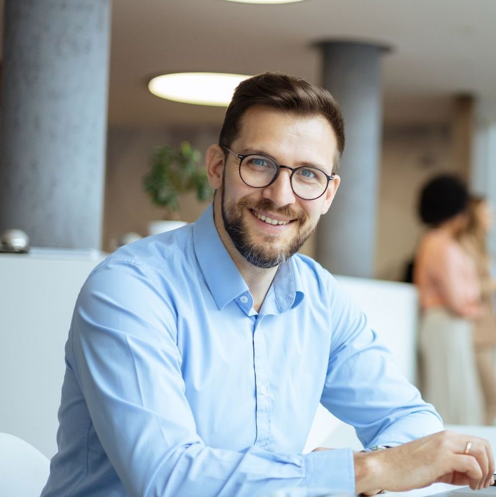 Man with glasses smiling, wearing a blue shirt in a bright office.