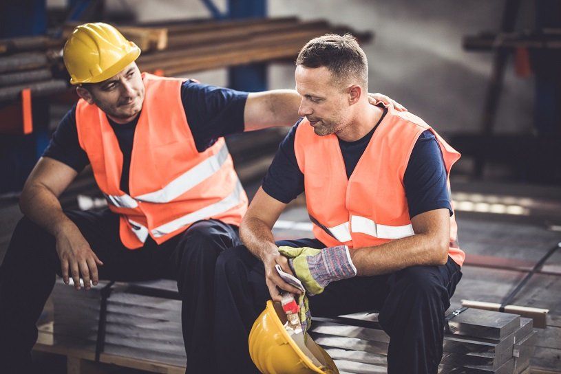 Two workers in safety vests; one comforts the other, sitting in a warehouse.