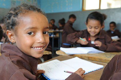 Girl smiling in a classroom, writing in a notebook, other students in background.