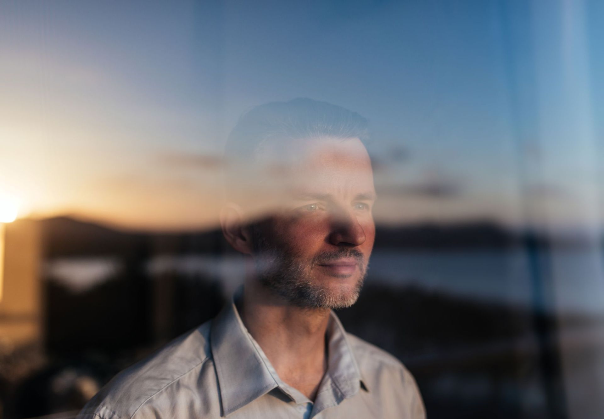 A man looking off-camera, reflected in a window against a sunset sky over water.
