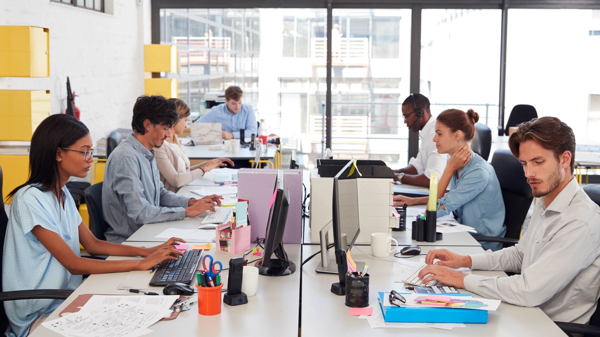 People working at desks in a bright open-plan office, using computers during a team meeting.