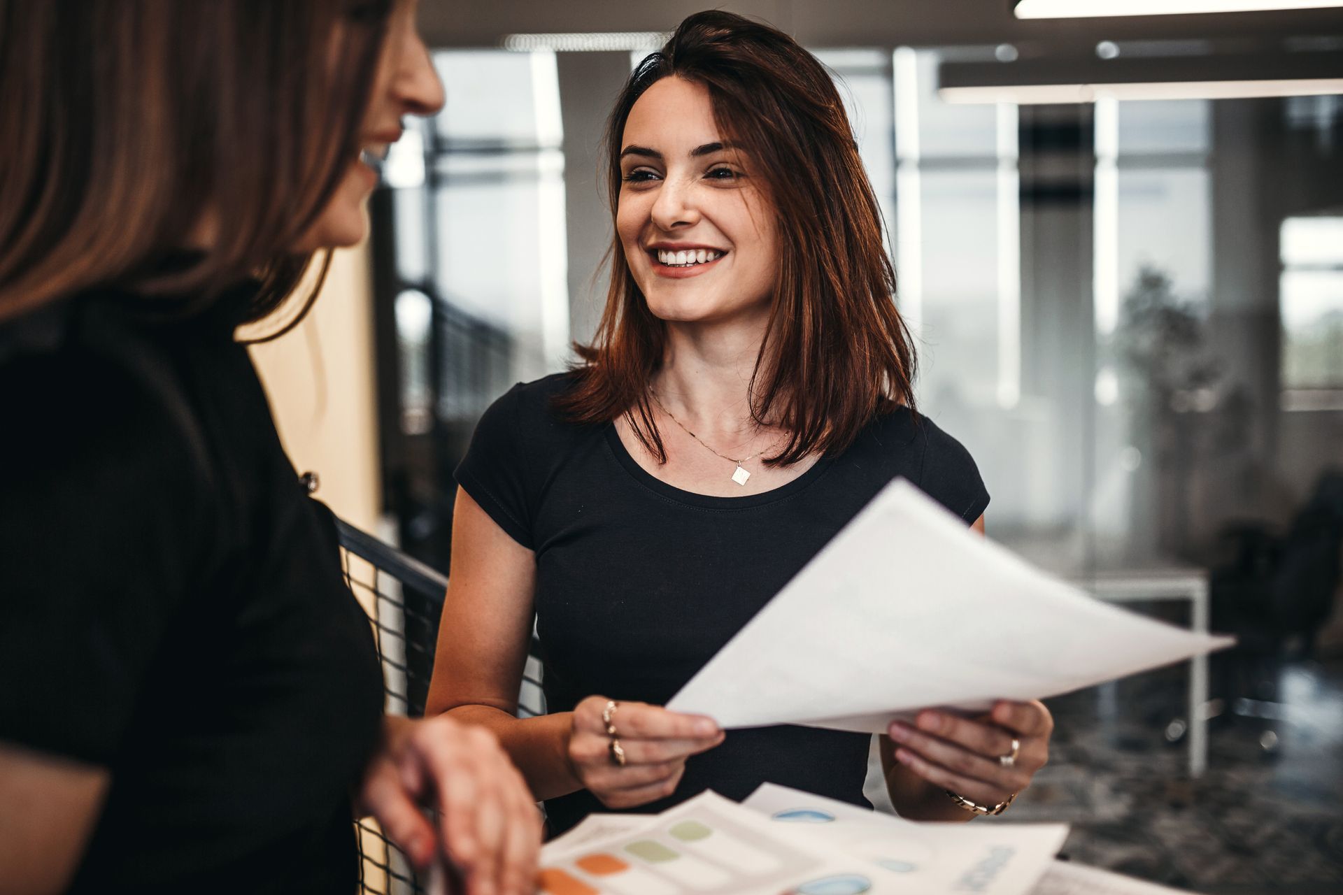 A professionally-dressed woman smiles as she hands a piece of paper to another woman.