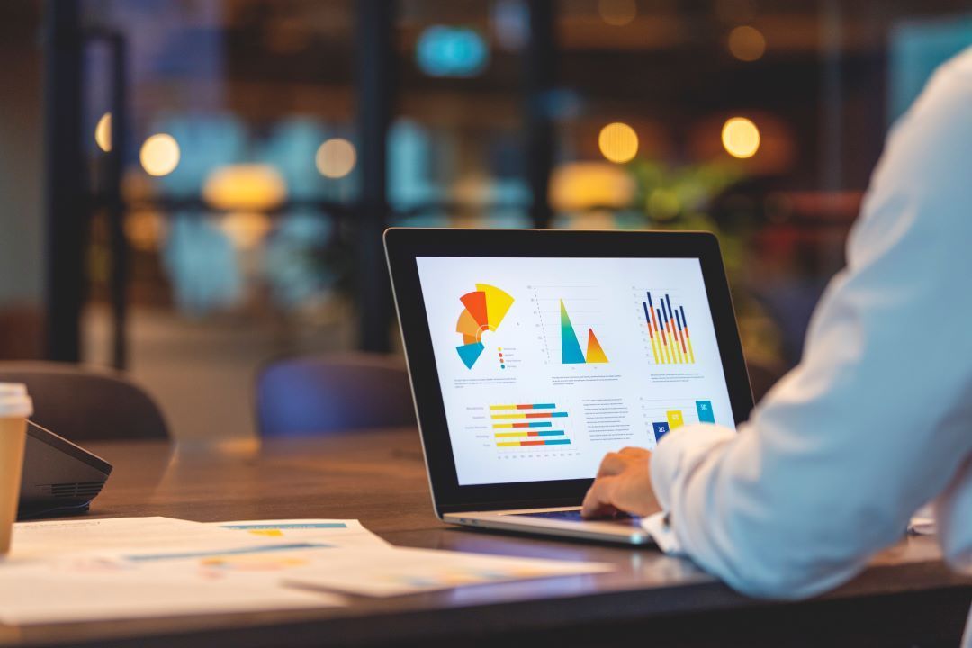 Person working on laptop, viewing graphs and charts at a desk, warm lighting, blurred background.