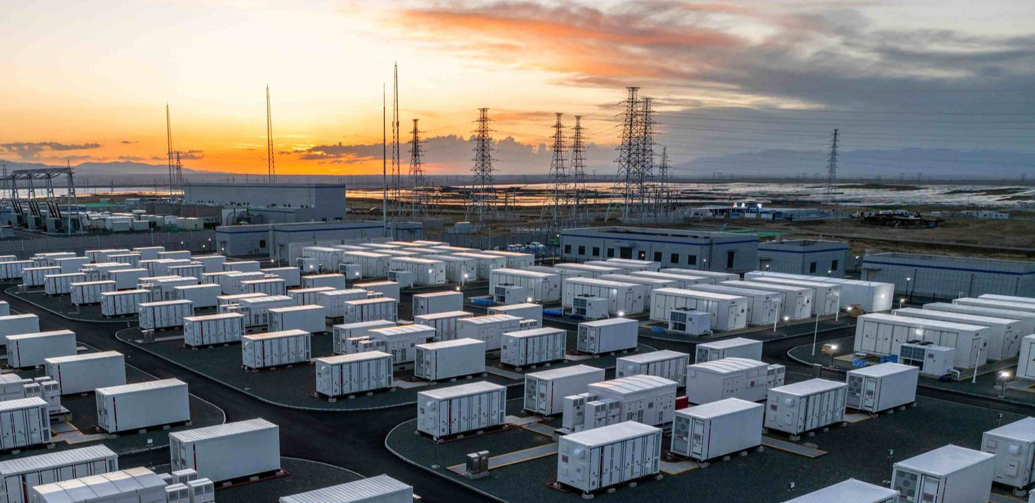 A large battery storage facility at sunset, with rows of white containers and power lines.
