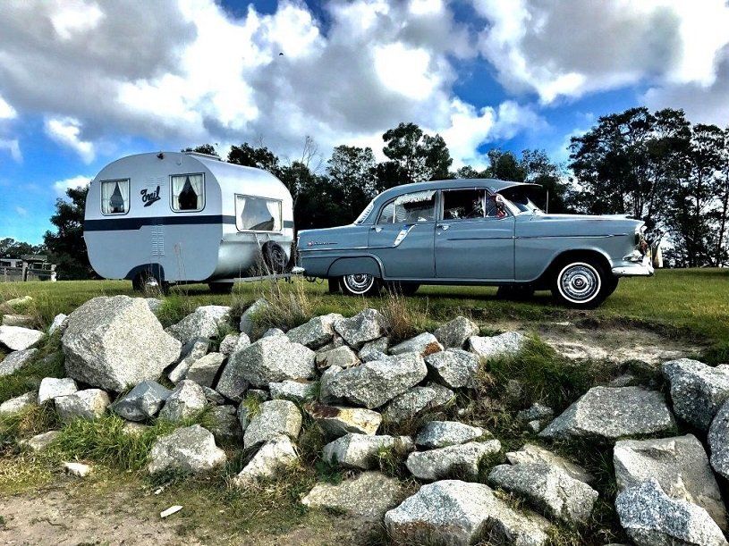 A classic gray car towing a vintage silver travel trailer on a grassy hill near rocks, under a cloudy sky.