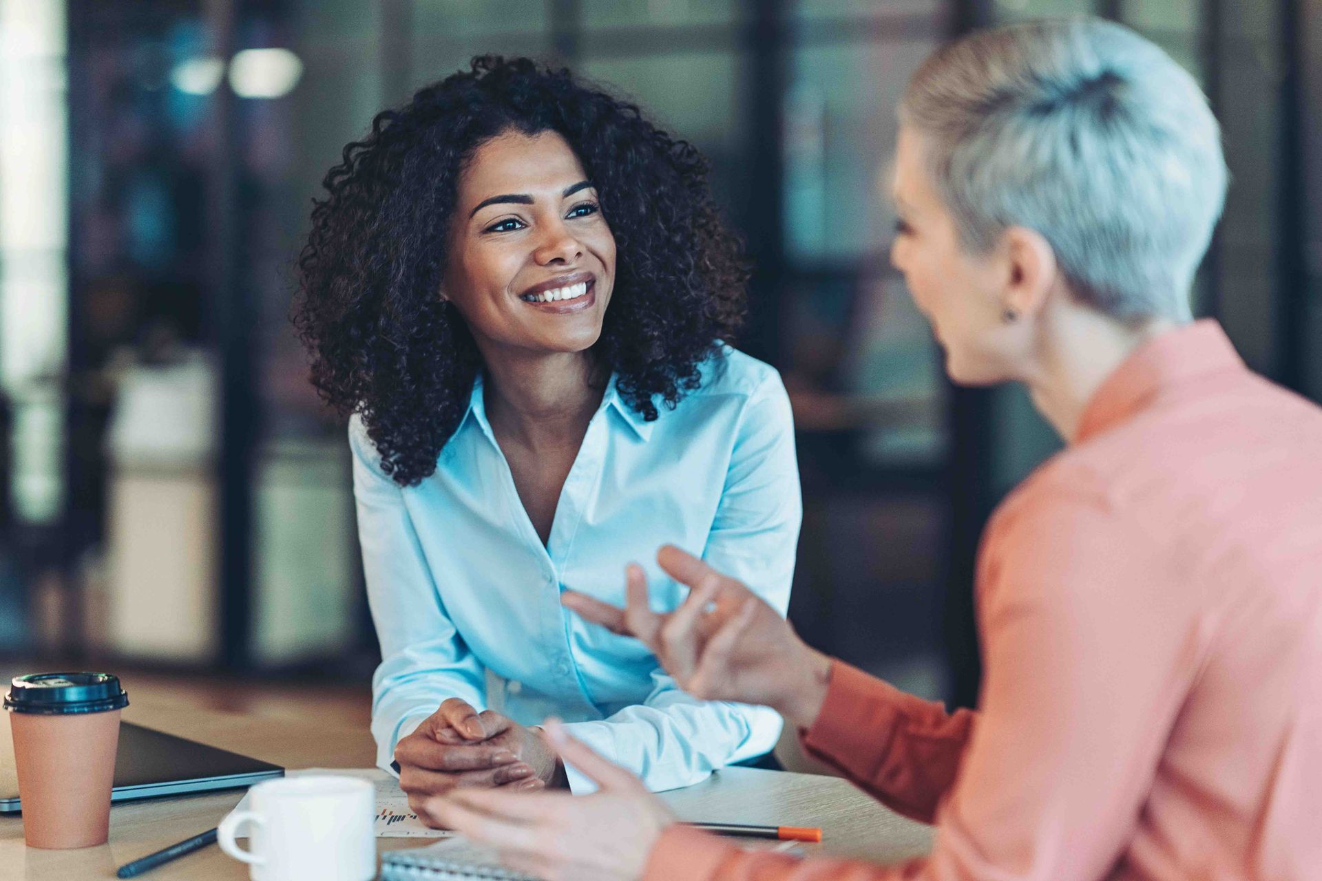 Two women in an office setting engaged in a discussion, one smiling while the other gestures.