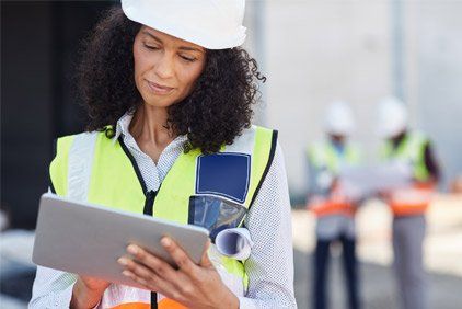 Woman in safety vest and hard hat using a tablet at a construction site.