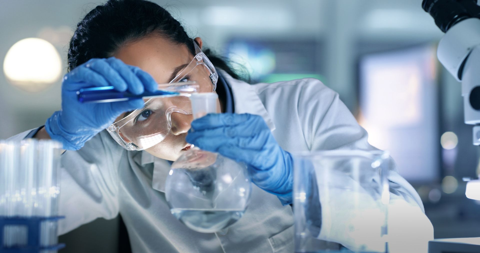 Scientist in lab coat and gloves, pouring blue liquid from a tube into a flask.