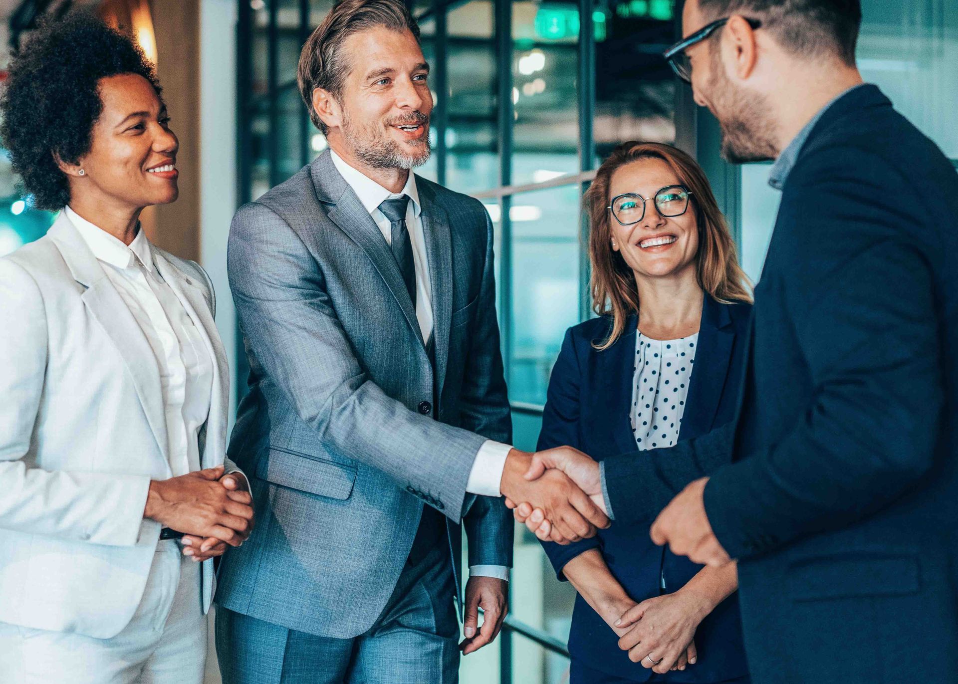 Four businesspeople in suits shake hands indoors, smiling.