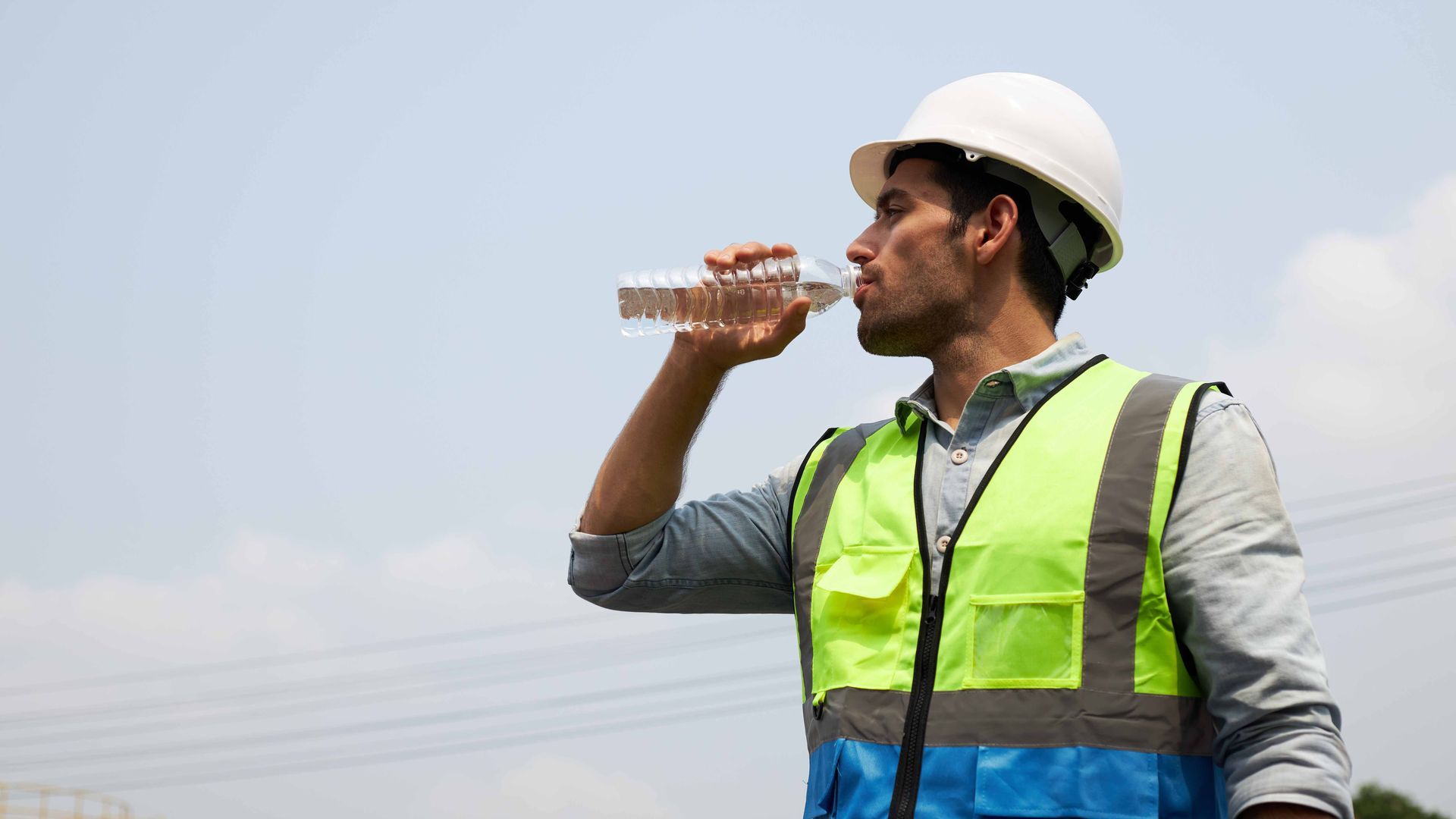Construction worker in safety vest and hard hat drinks water outdoors.