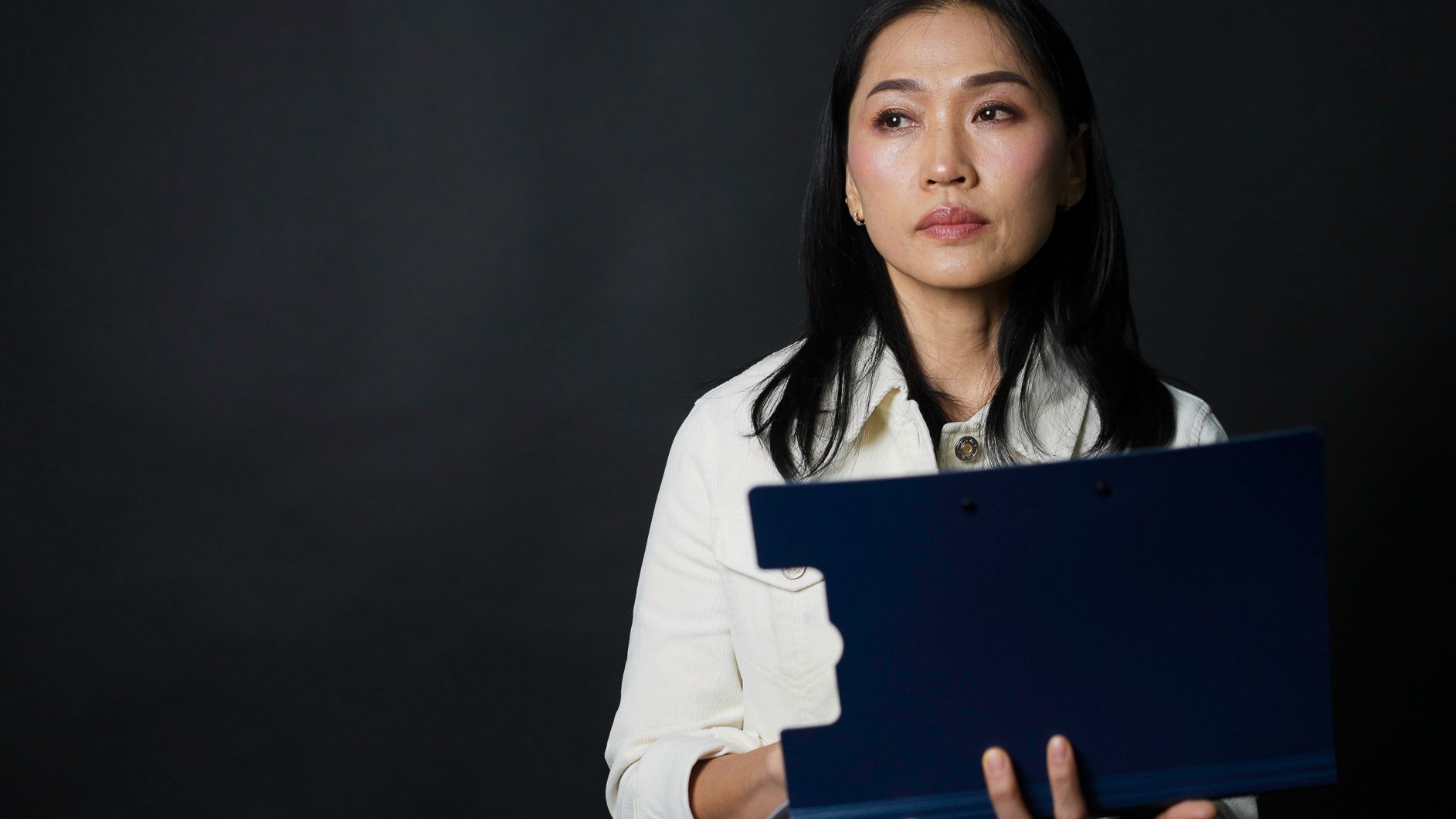 Woman in white jacket holds a blue clipboard, looking thoughtful, against a dark background.