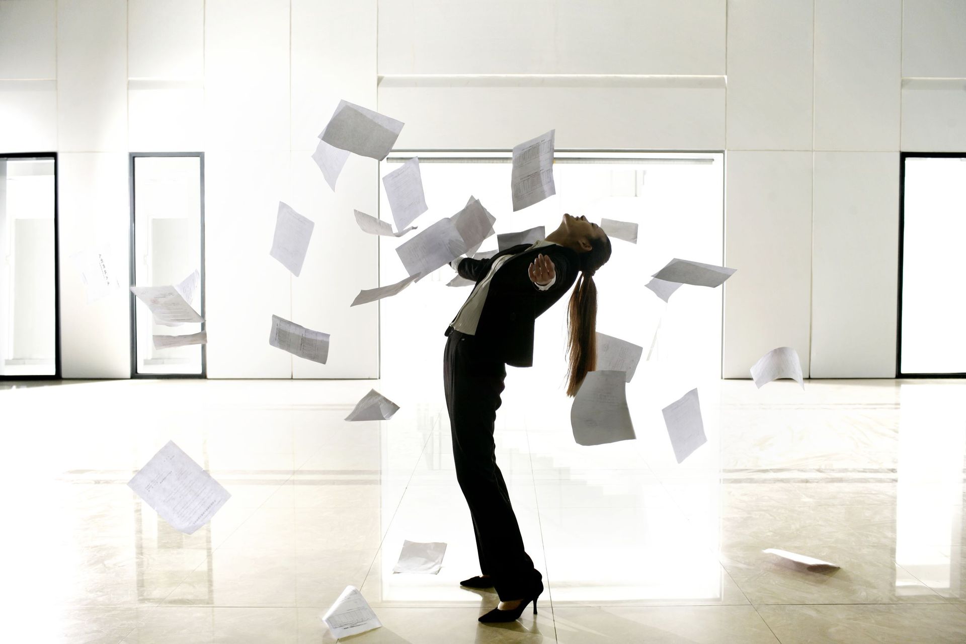 Woman in a suit, head back, surrounded by papers flying in air, in a modern, light-filled room.