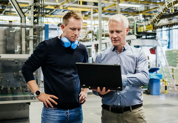 Two men looking at a laptop in a factory; one with headphones, the other pointing.