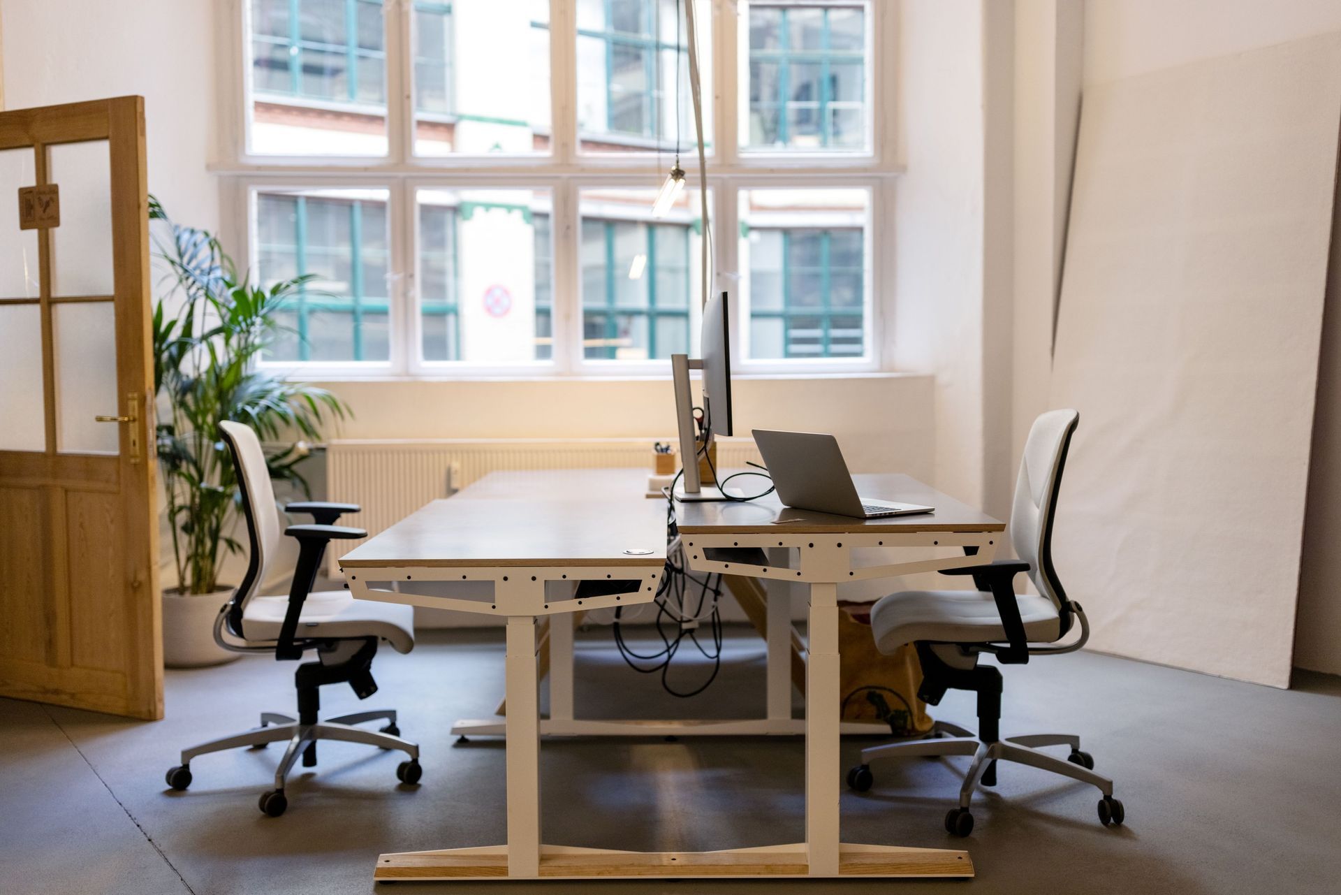 Bright office with wooden desks, chairs, laptops, and large windows with plants nearby