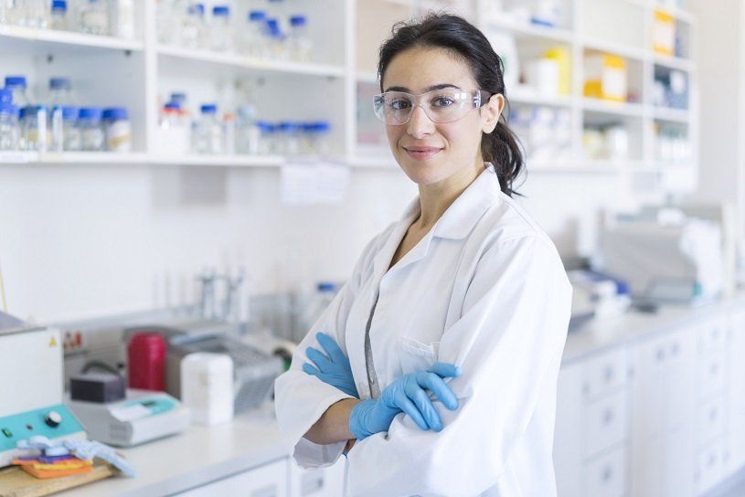 Scientist in lab coat, safety glasses, and gloves, arms crossed, smiling in a laboratory setting.