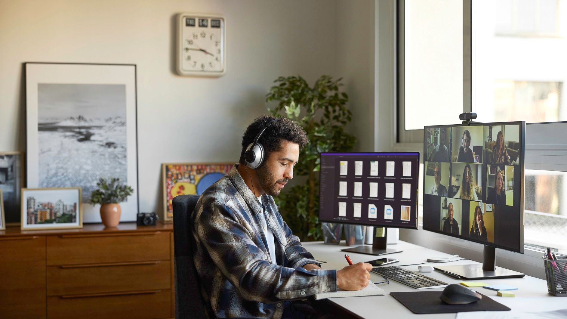 Man at a desk wearing headphones, working on a computer in a sunlit home office.