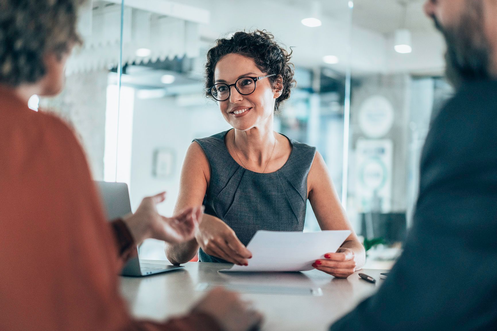 Woman in glasses reviews document with two people at table in office.