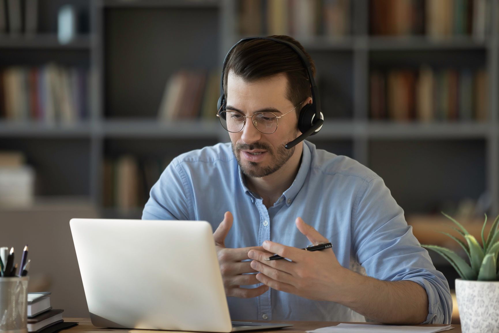 Man wearing a headset, gesturing during a video call on his laptop. Bookshelves in background.