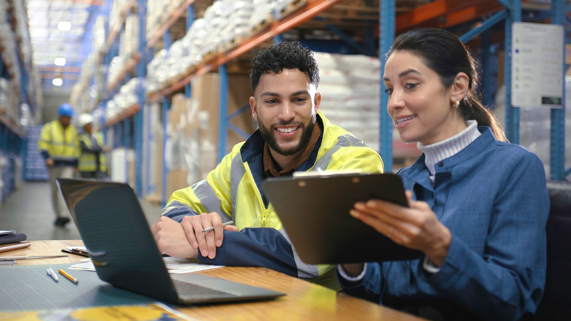 Two people reviewing documents and laptop in a warehouse. Man in safety vest smiles, woman points.