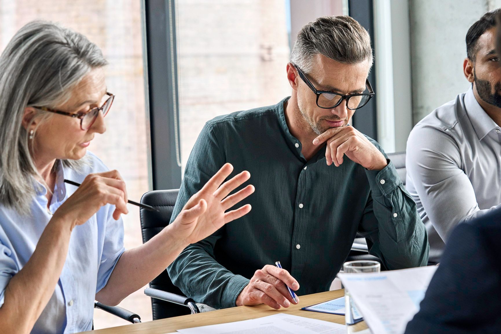 People in a business meeting around a table, one woman gesturing, one man pondering.