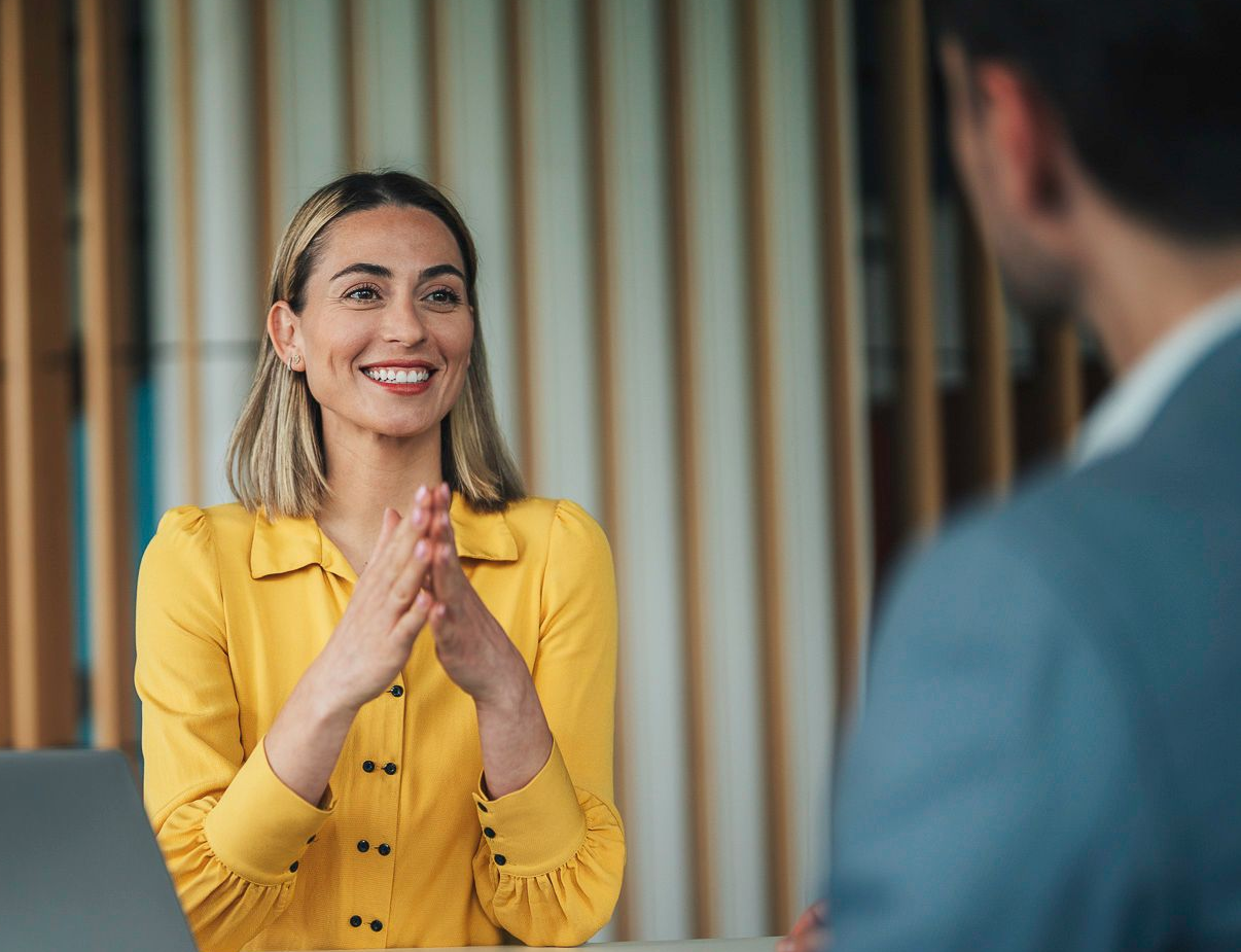 Woman in yellow shirt, smiling, clapping hands, facing a person in a suit, in an office setting.