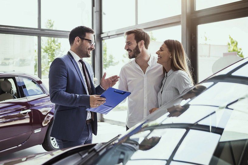 Car salesman showing paperwork to a couple at a dealership, cars visible in background.