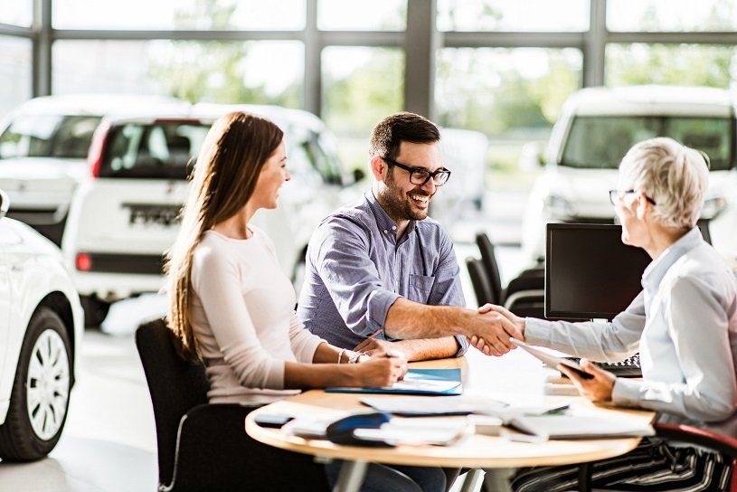 Couple shakes hands with salesperson in a car dealership, cars in background.