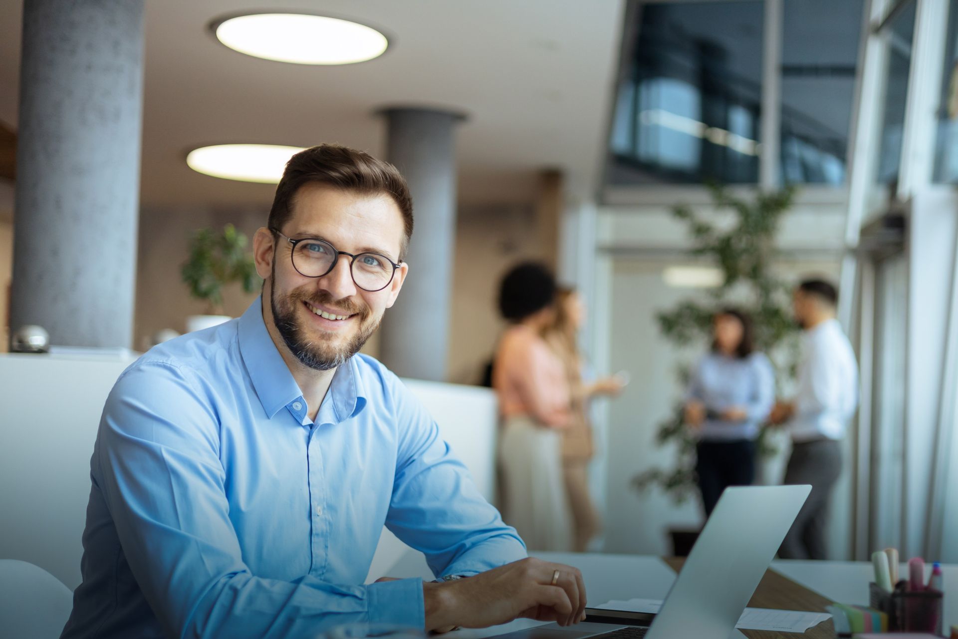 Man with glasses, smiling, working on laptop in modern office, colleagues in background.