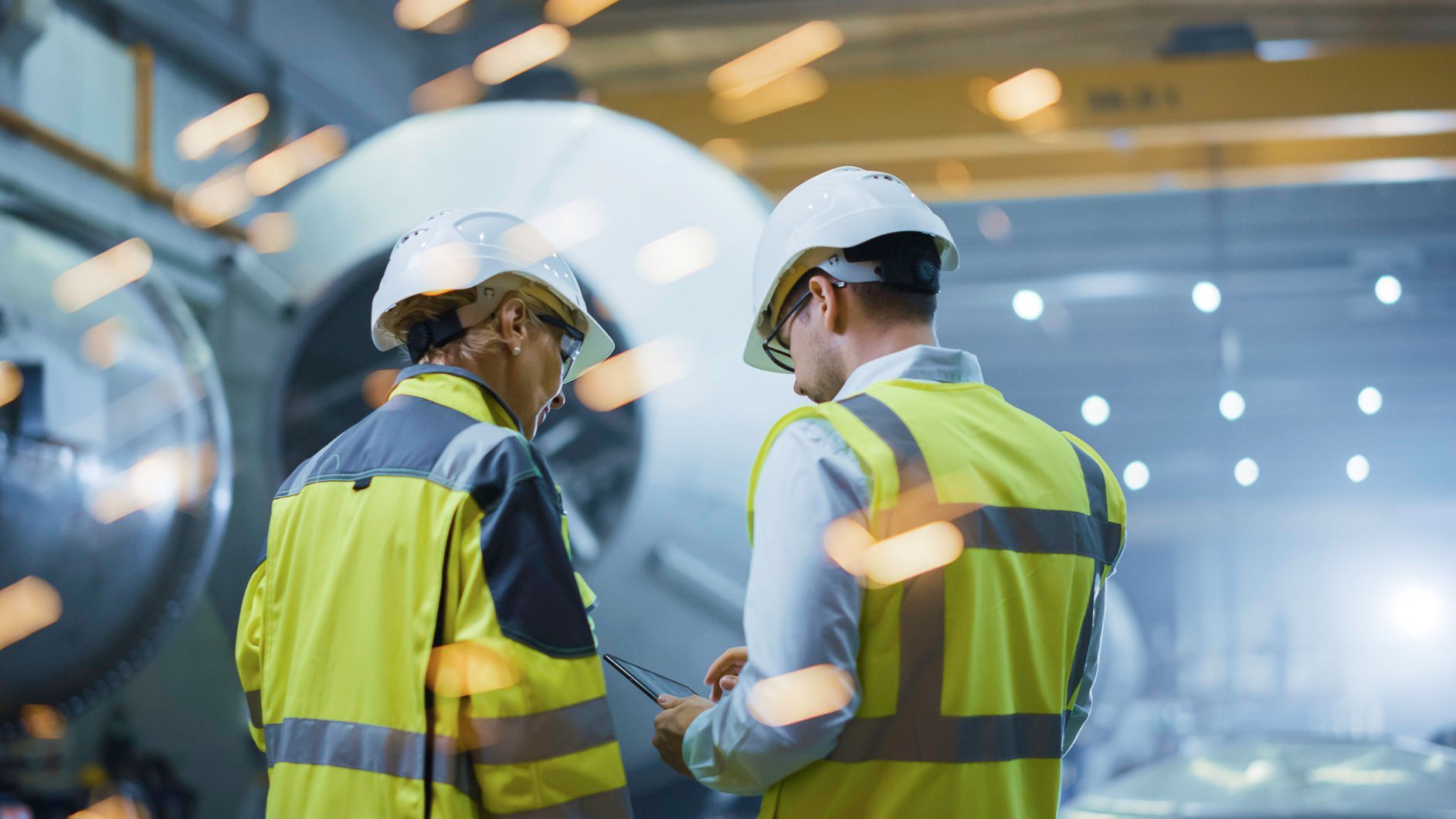 Two engineers in safety vests and hard hats discussing something near machinery in a factory.
