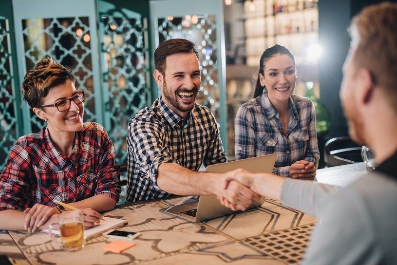 People at table shake hands, smiling. Restaurant setting. Laptop, notes, and drink present.