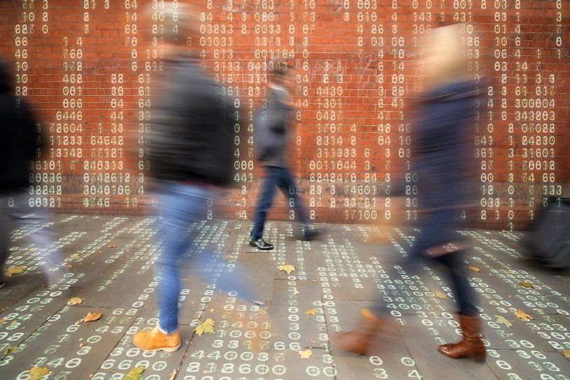 Blurred figures walk past a wall with projected binary code.