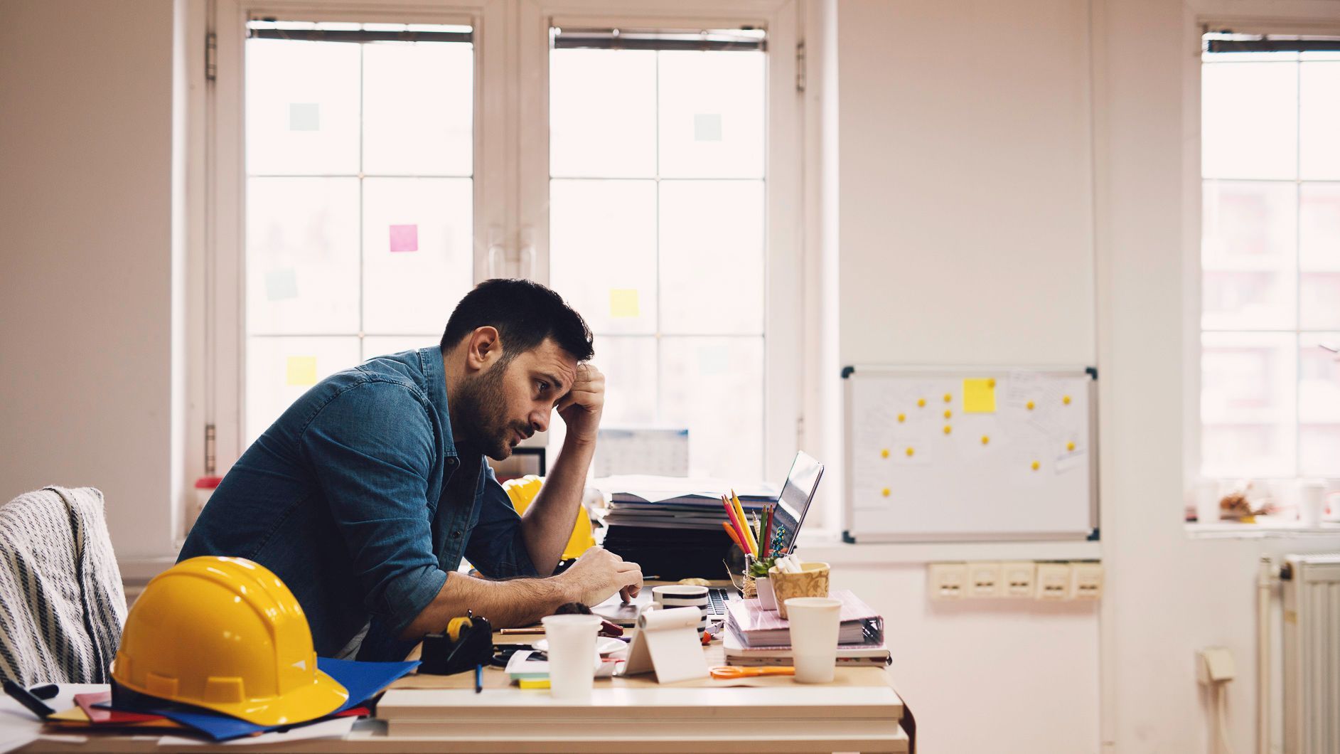 Man leaning over a desk in an office, looking stressed amid papers, a laptop, and a yellow hard hat.