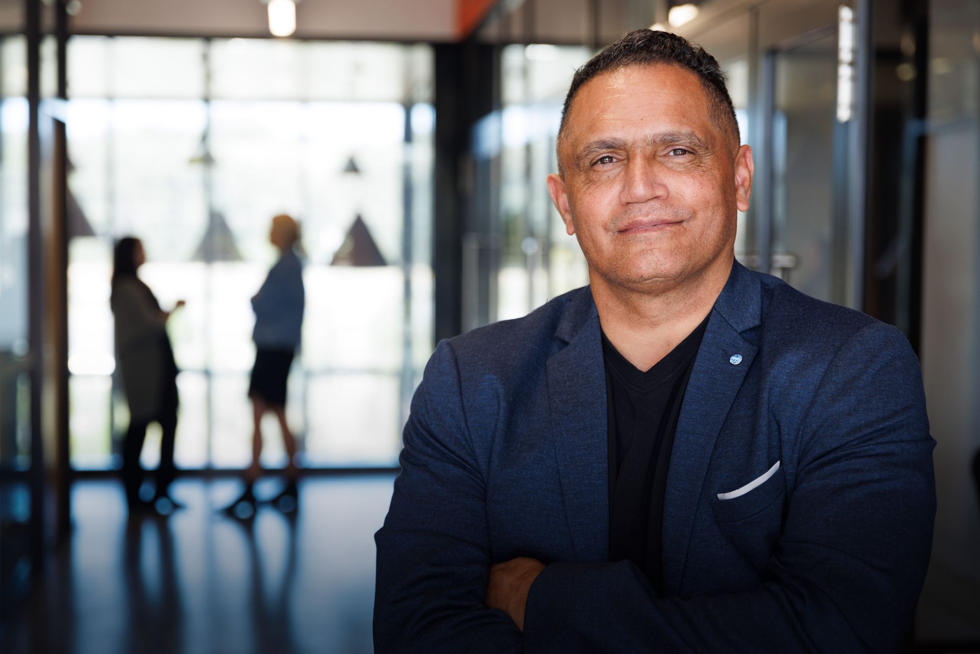 Man in blue blazer, arms crossed, smiles in office setting with figures blurred in background.