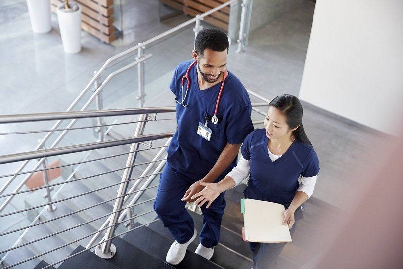 Two medical professionals in blue scrubs walk up stairs, one with a hand on the other's arm, inside a building.