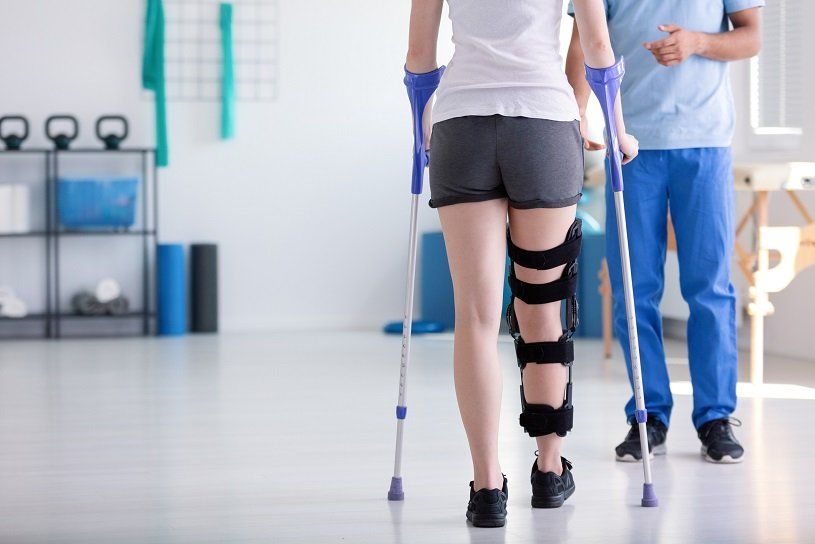 Woman with knee brace using crutches, assisted by a person, in a rehabilitation setting.