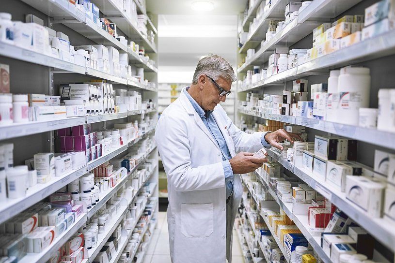 Pharmacist in a white coat looking at medication on shelves in a pharmacy aisle.