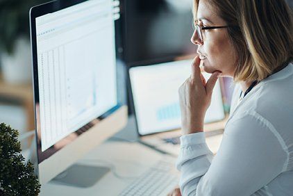 Woman with glasses thoughtfully examines a computer screen displaying graphs, a laptop in the background.